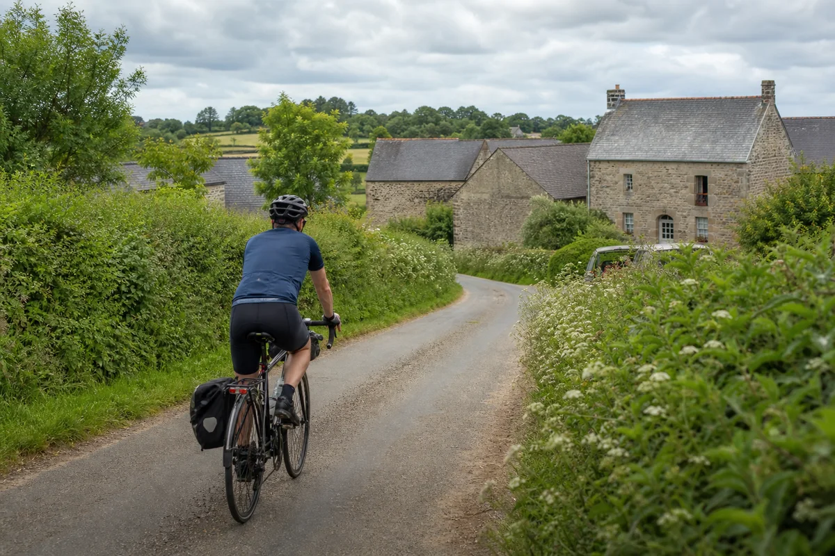 A cyclist riding a quiet lane between hedges and stone farm buildings